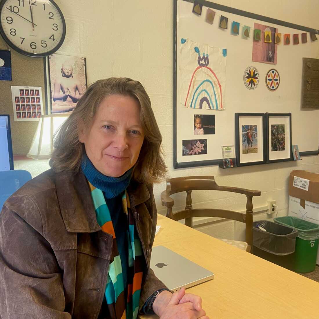 In this photo, Mariana Chilton, a professor of public health at the University of Massachusetts Amherst, sits at a table with a closed laptop computer on it. On the wall behind her are a whiteboard and clock.