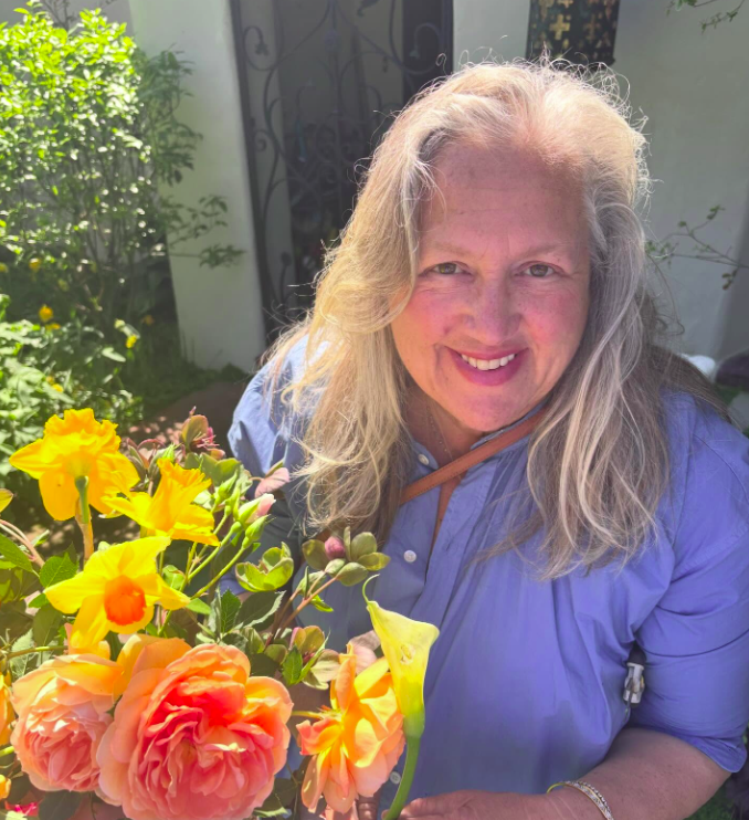 A woman with a light skin tone smiles while holding colorful flowers including yellow daffodils and orange roses in an outdoor garden setting.