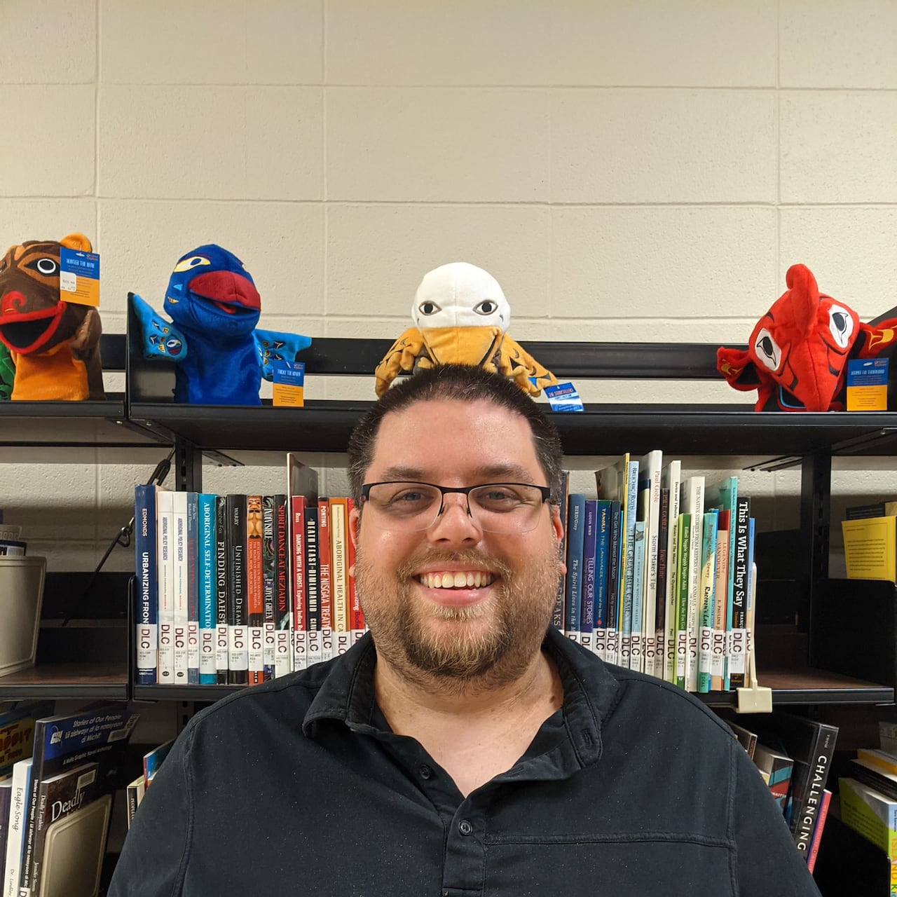 A man with glasses wearing a black shirt stands before a bookshelf. There are some stuffed creatures on top of the bookshelf.