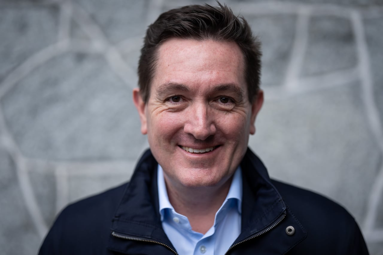A white man smiles in front of a grey background.
