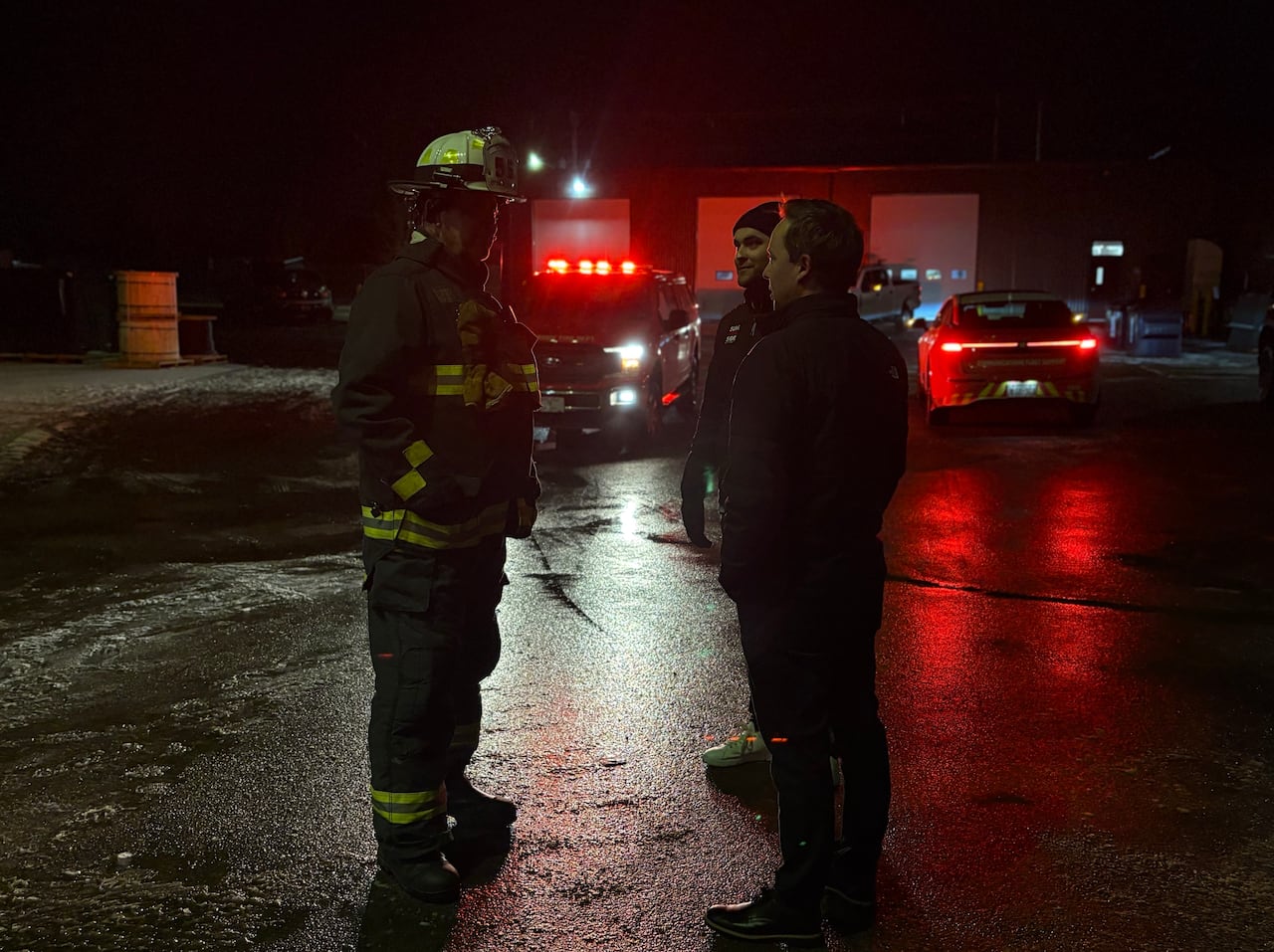 A firefighter stands across from two men at night, with the red lights of emergency vehicles visible in the background.
