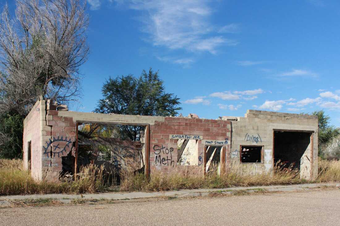 A cinder-block structure has no roof, and graffiti that says "Stop Meth" is written on the wall. Tall grass surrounds it, and a blue sky is visible.