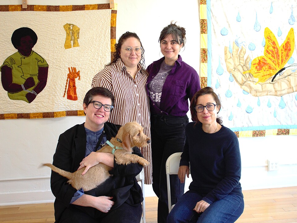 Four women pose in a group portrait. One of them holds a small yellow dog. Behind them are two quilts hanging in an art gallery.