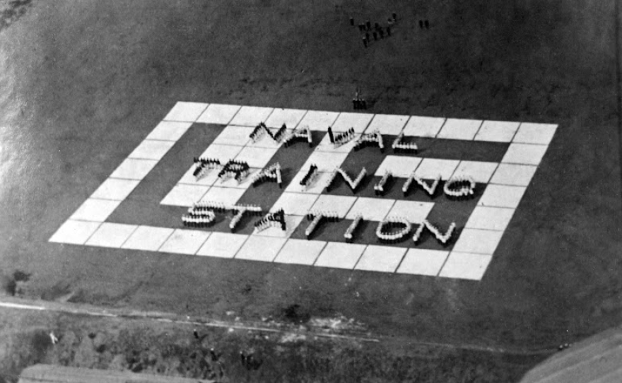 One b/w photographic print overlooking the parade grounds at the U.S. Naval Training Center in San Diego in about 1925. On the grounds, naval recruits are arraigned to spell out "Naval Training Station."
