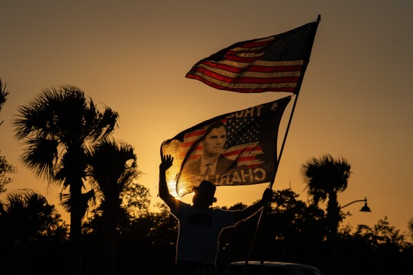 President Trump supporter Steve Emery holds a flag paying tribute to right-wing political activist Charlie Kirk along a bridge to Mar-A-Lago before President Donald Trump departs West Palm Beach, Fla., for Washington, Sunday, Jan. 4, 2026. (AP Photo/Allison Robbert, File)