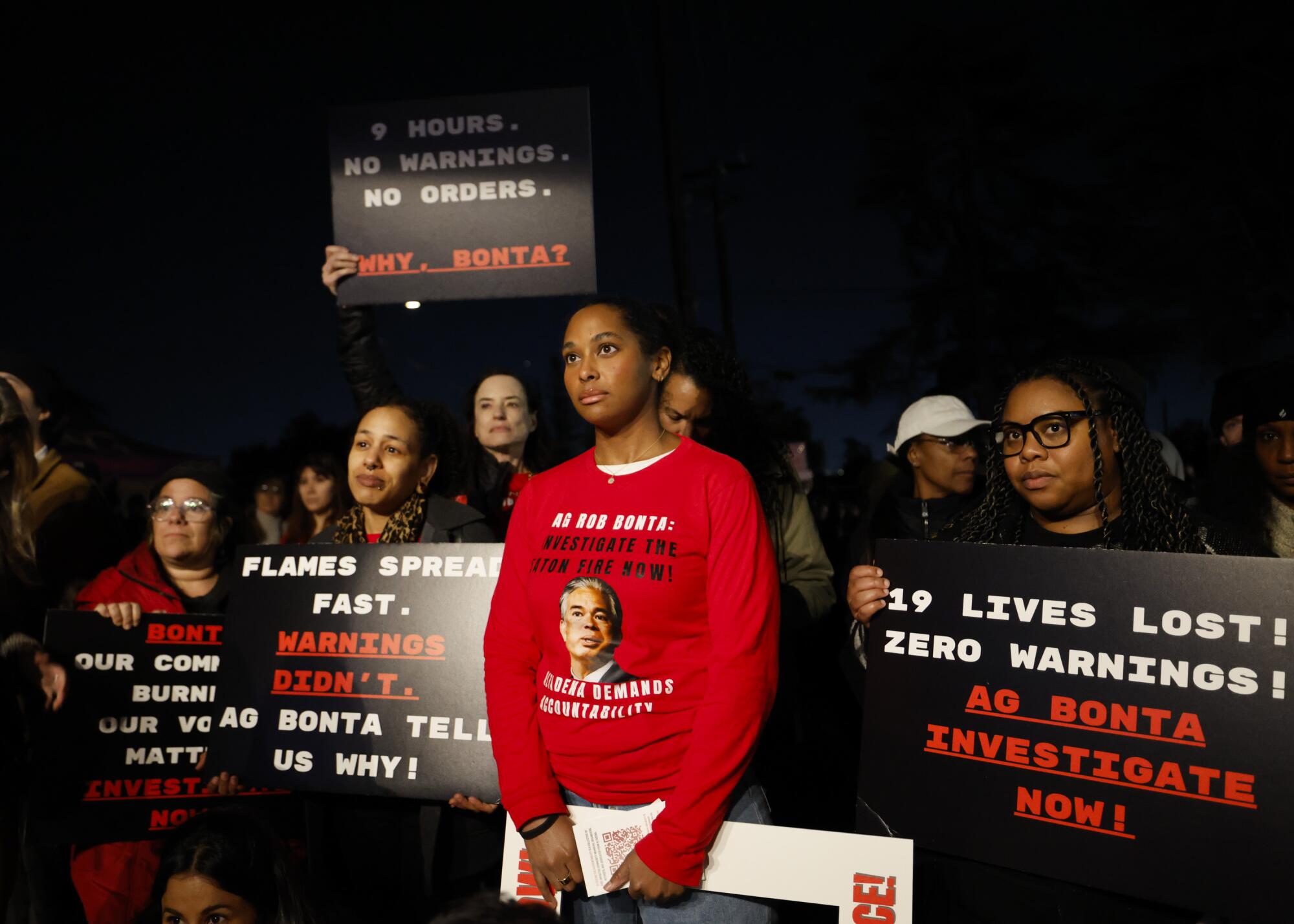 People stand together and hold signs at a protest gathering. One wears a shirt with Rob Bonta's face on it.