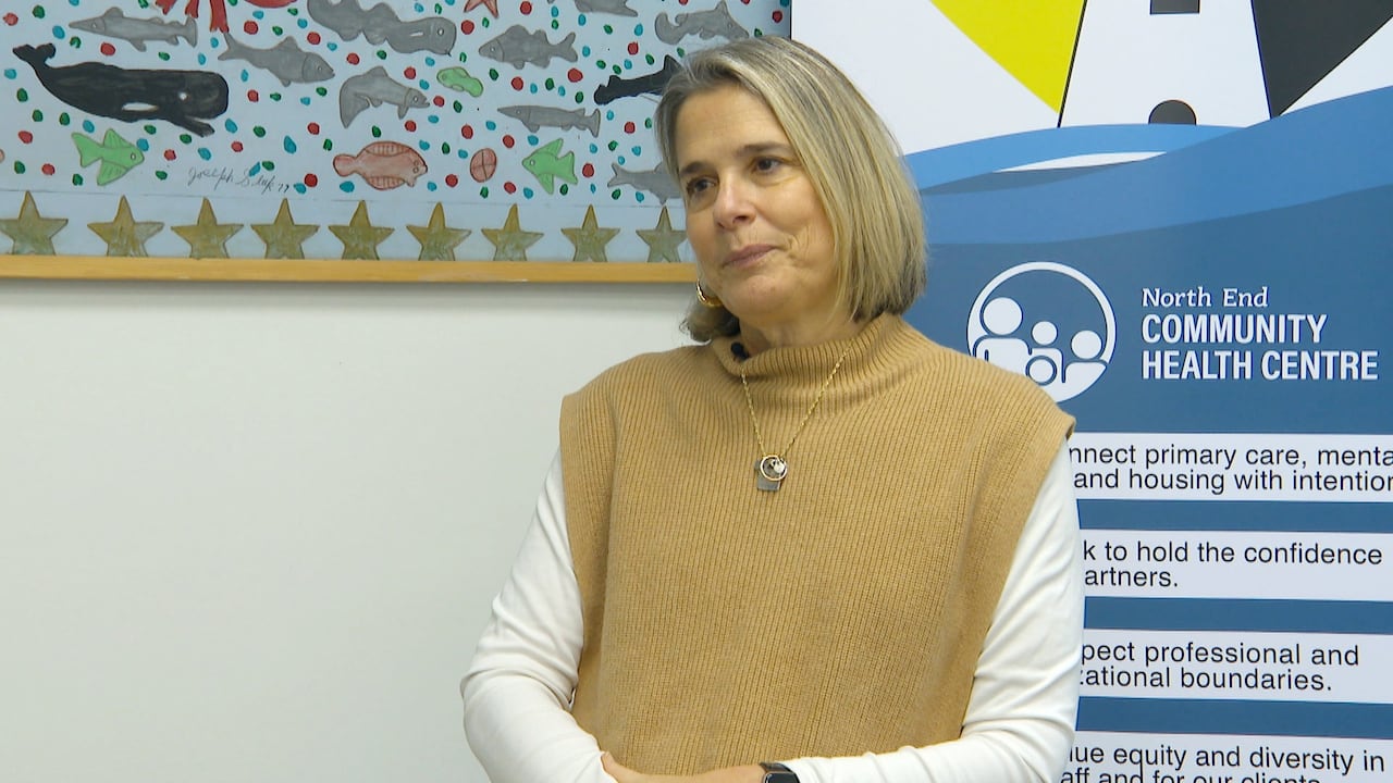 A white woman with a brown sleeveless sweater over a white blouse sits in an office in front of a sign for the health centre