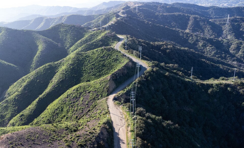 An aerial view of a road traversing the ridgeline of mountainous area. Greenery on both sides with power poles running down the middle. 