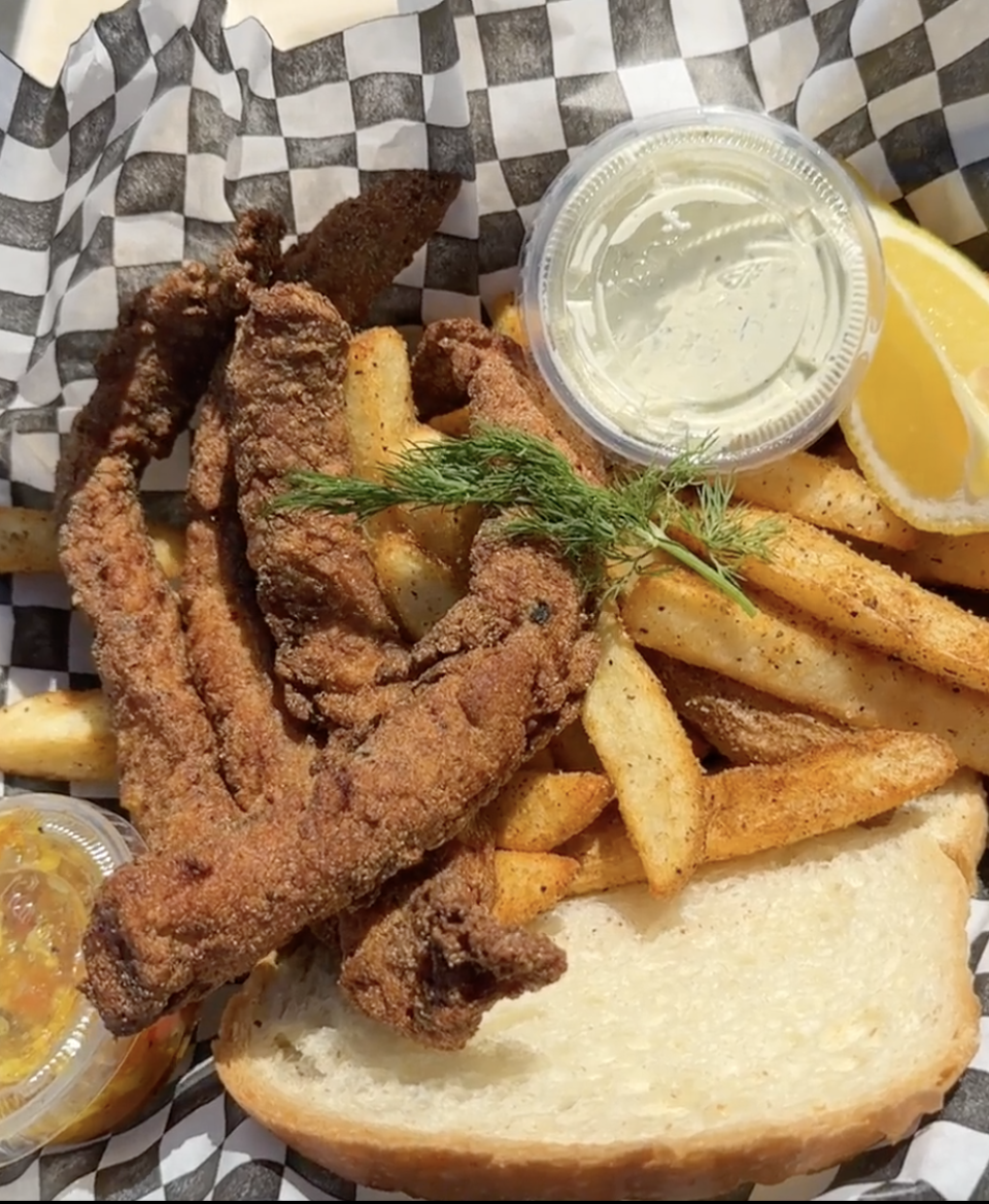 A basket lined with black and white checkered paper holds golden-brown fried fish filets, thick-cut French fries, a slice of white bread, a lemon wedge, fresh dill garnish, and two small containers of sauce