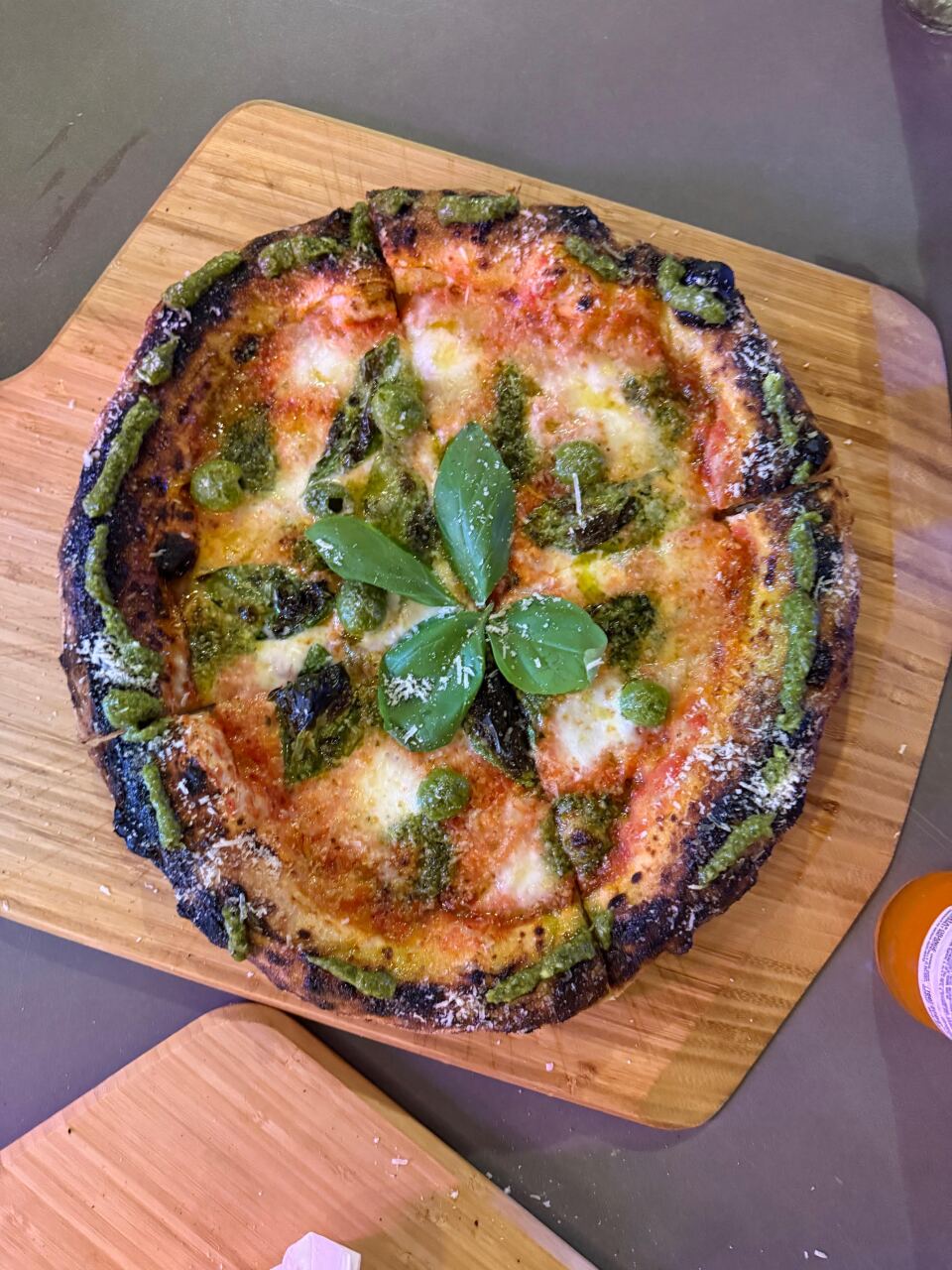 A charred Neapolitan-style pizza on a wooden cutting board topped with melted mozzarella, green pesto or herb sauce drizzled in a pattern, and fresh basil leaves in the center