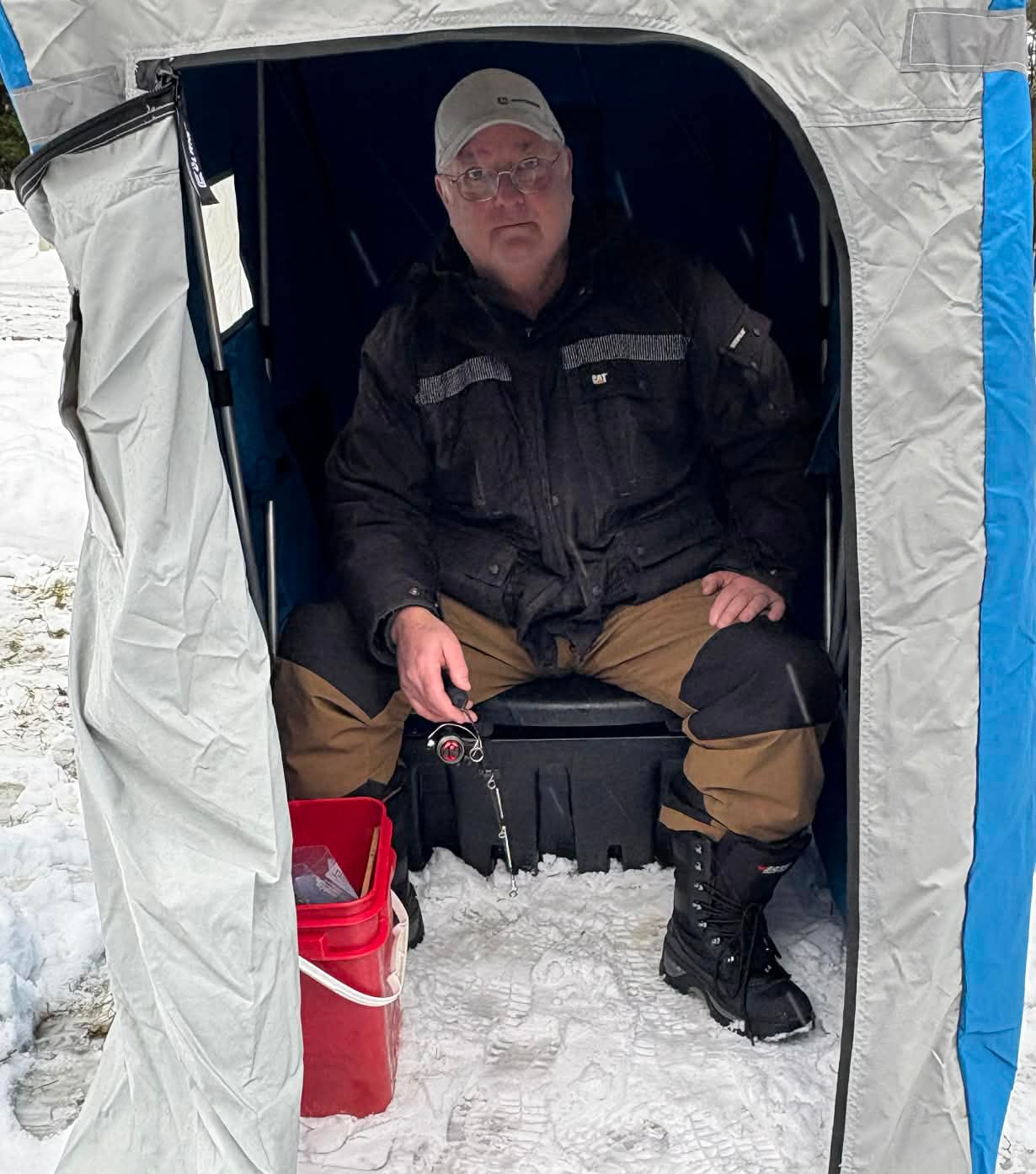 A man with glasses and a white baseball cap fishes from inside a small tent on the ice.