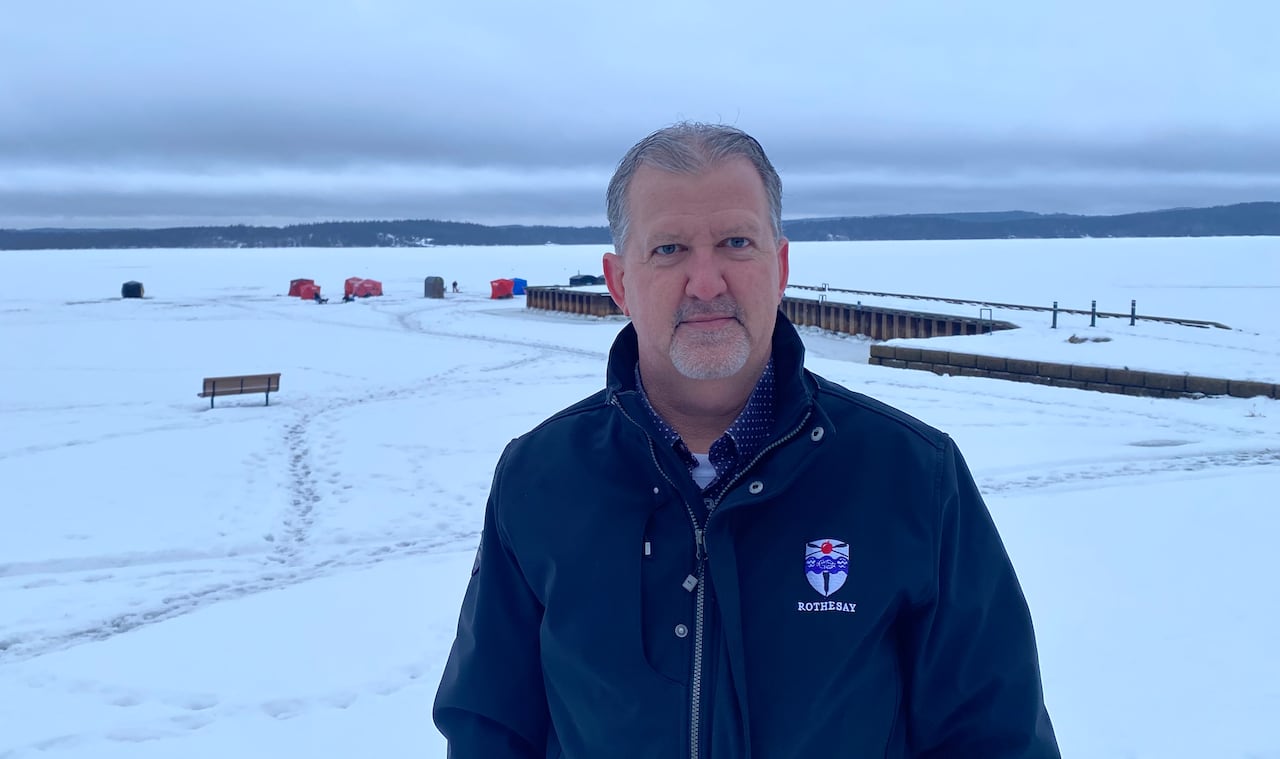 A man in a blue jacket stands in front of an open river, with a wharf and small ice-fishing village in the background in the background. 