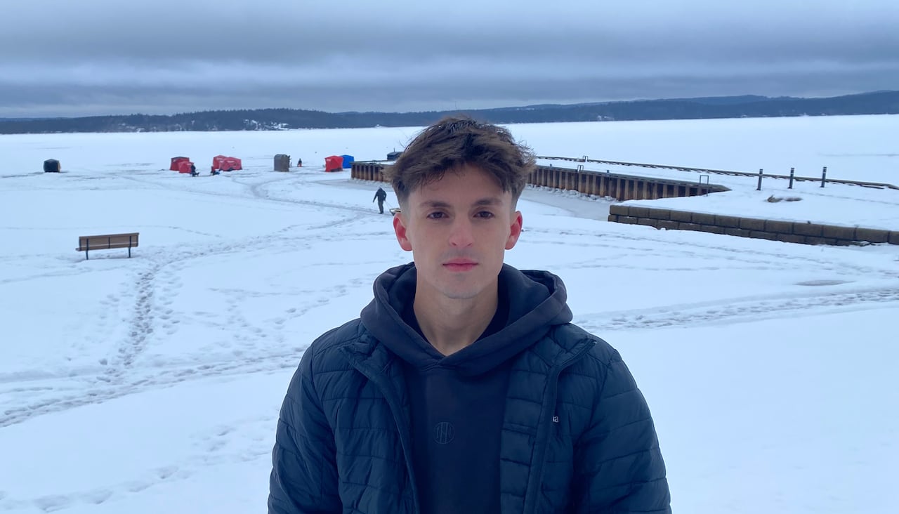 A man with dark hair and a black jacket stands in front of an open river with a wharf and small ice-fishing village in the background.
