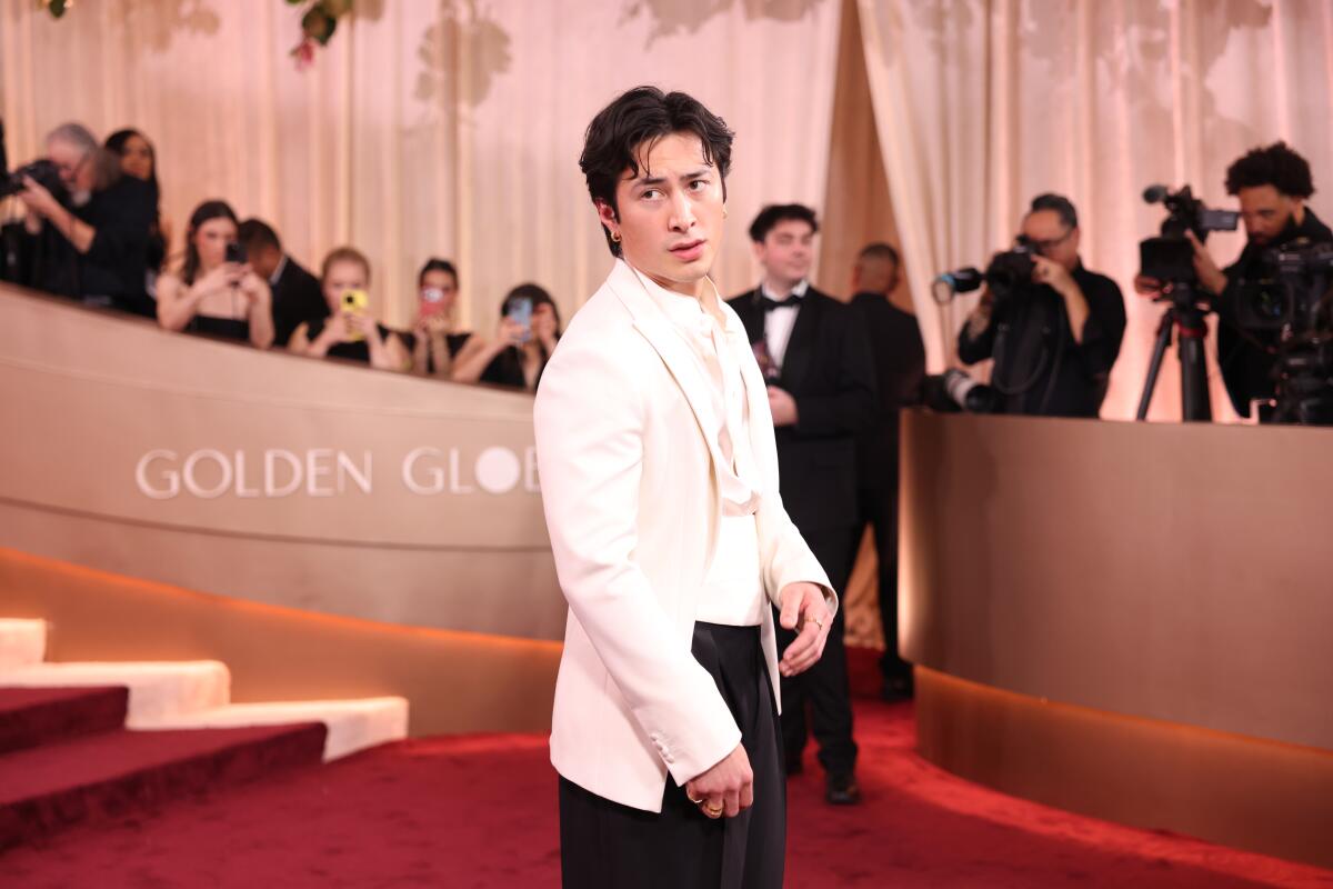 Hudson Williams, wearing a white tux jacket, looks over his right shoulder on a red carpet