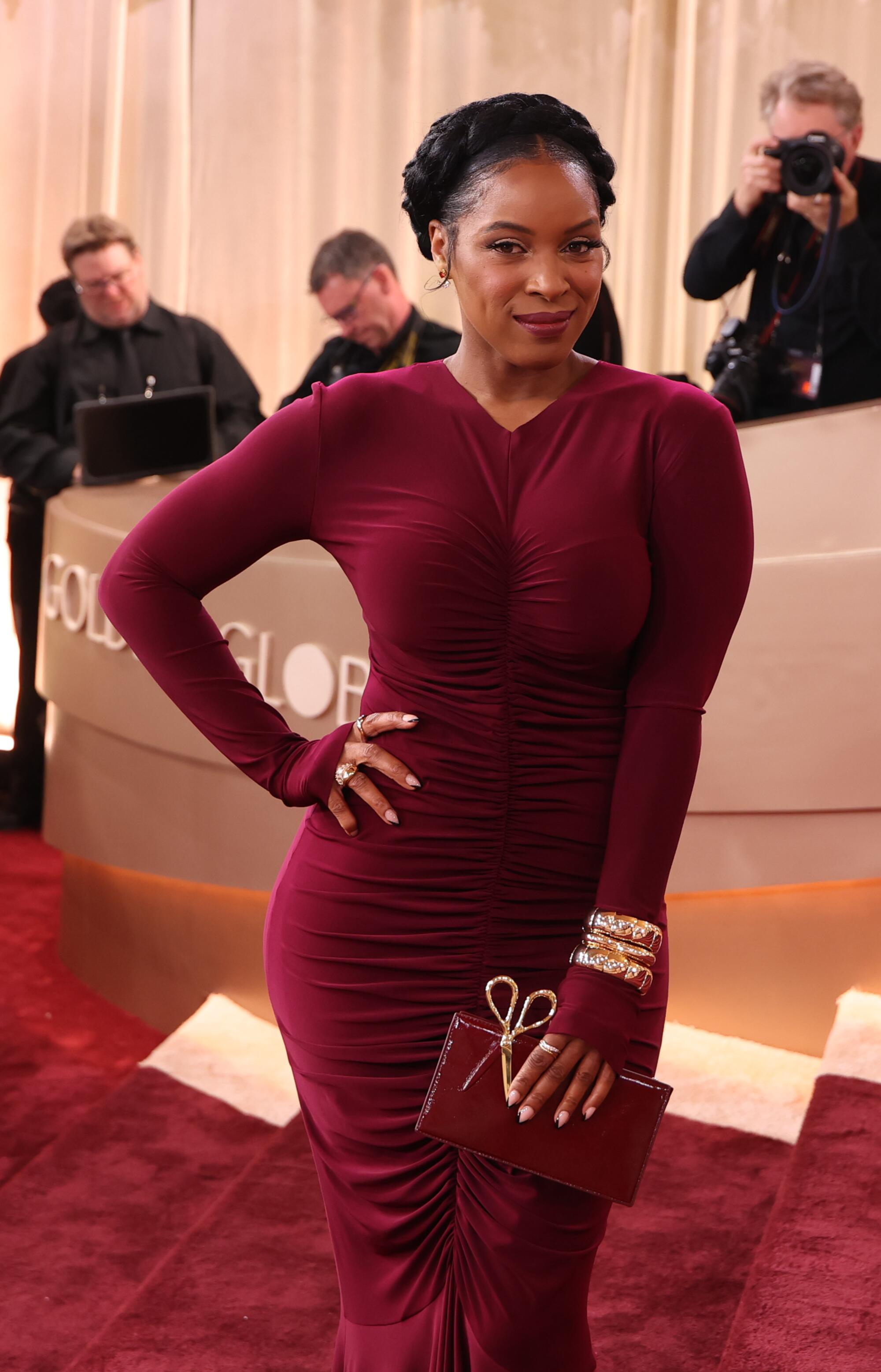 Janine Rubenstein, wearing a burgundy form-fitting dress, walks the red carpet. 