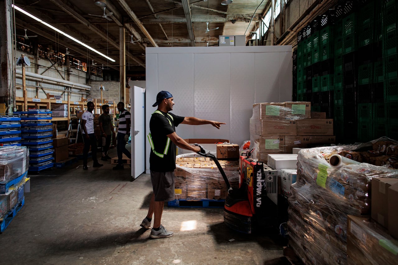 A man wearing black shorts, a black T-shirt and a backwards hat moves boxes full of food around a warehouse.