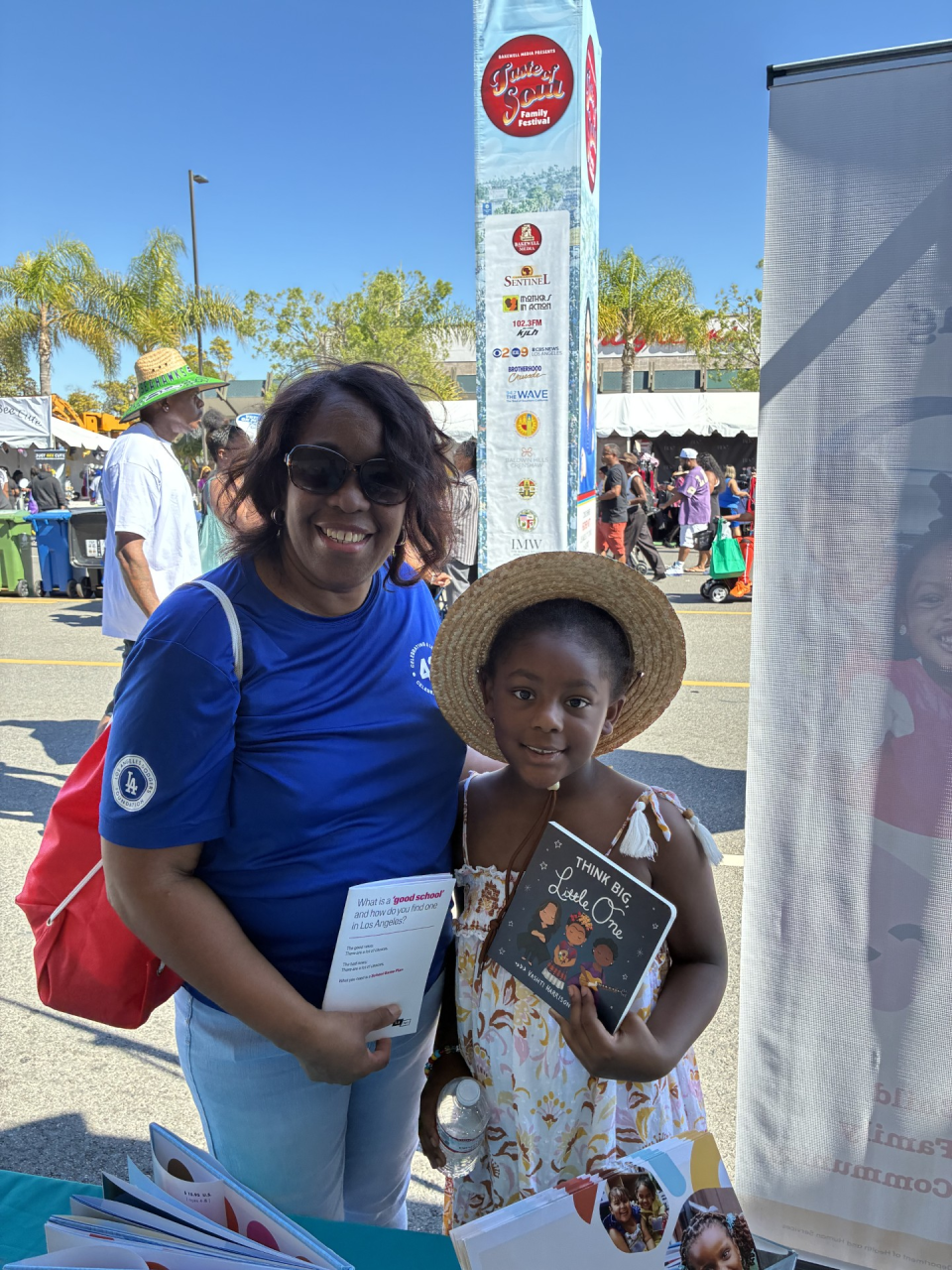 An adult with medium-dark brown skin and a young child with medium-dark brown skin smile at an outdoor community festival. The adult wears a blue T-shirt and sunglasses and holds a pamphlet about finding a good school in Los Angeles. The child wears a straw hat and a patterned dress and holds a children’s book titled Think Big, Little One. They stand behind an information table with flyers, with festival tents, banners, and people in the background under a clear blue sky.