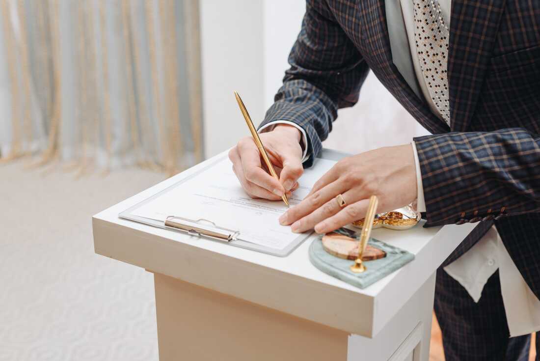 A young man standing at wedding ceremony and signing wedding documents, groom in black costume, cropped, unrecognizable.