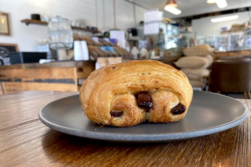 A close up of a luscious chocolate croissant, sitting on a grey plate on a wooden table in a bakery cafe