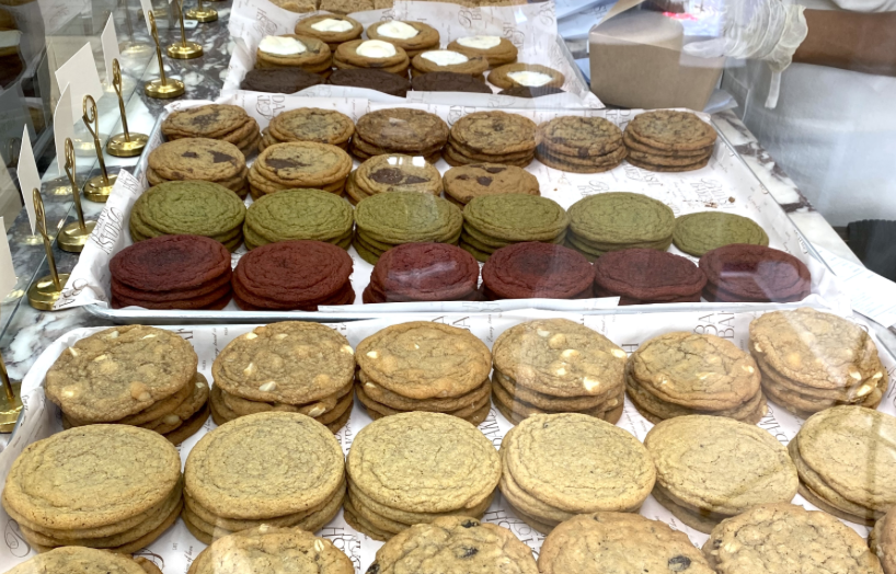 Rows of perfectly round cookies of different colors (chocolate, cream, matcha) line up on a counter behind a piece of glass 