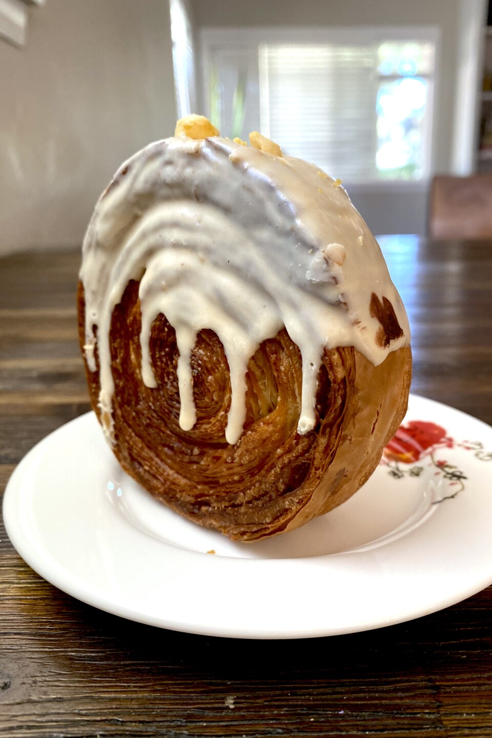A spiral croissant is standing on its side, covered in luscious white icing that's dripping down. It's on a white floral plate, sitting on a wooden table