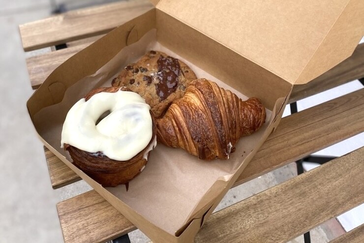 An open take out brown box contains three pastries; a cinnamon roll with a large swathe of white icing; a croissant, and a chocolate chip cookies. The box sits on a wooden table with slats.
