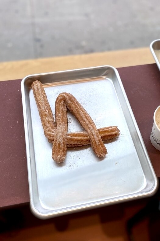 Two churros which have been laid out to spell out LA, sit on a metal tray, on a wooden table