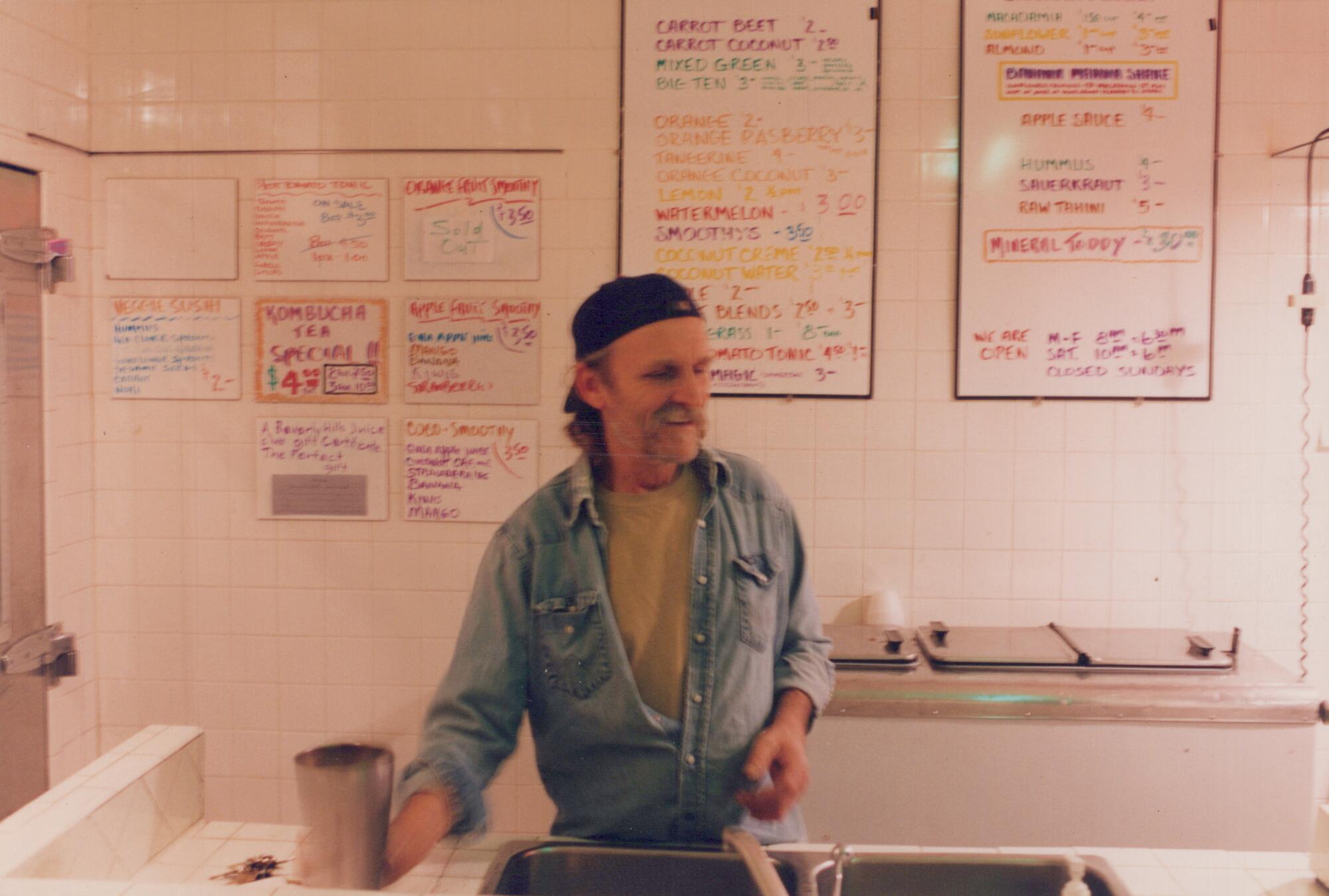 A man in a chambray shirt with a backwards baseball cap stands behind a counter