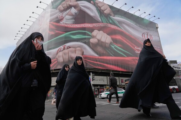Women cross a street under a huge banner showing hands firmly holding Iranian flags as a sign of patriotism, as one of them flashes the victory sign, in Tehran, Iran, Wednesday, Jan. 14, 2026. (AP Photo/Vahid Salemi)
