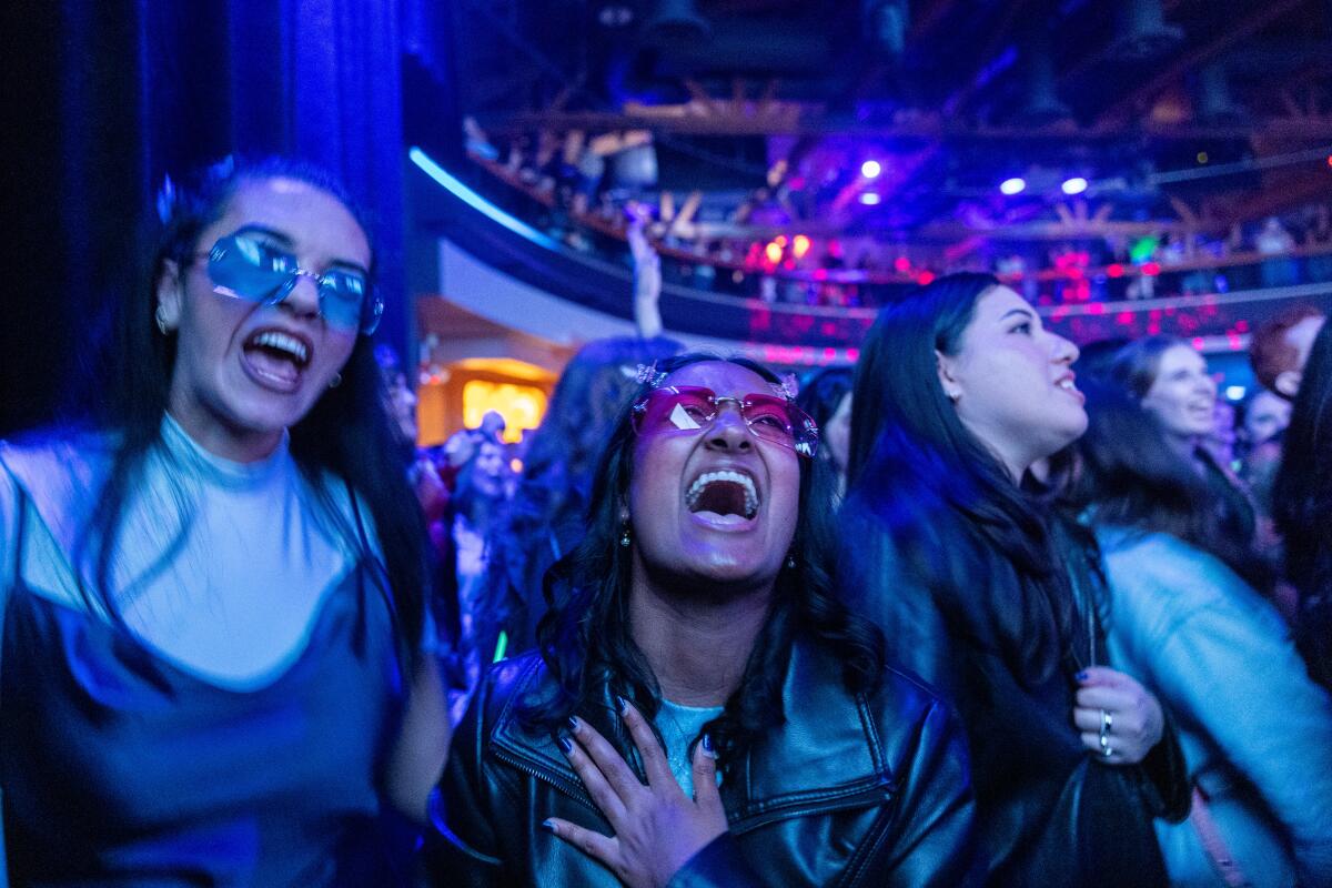 A a pair of women wearing colorfully tinted sunglasses scream as they stand on a crowded dance floor.