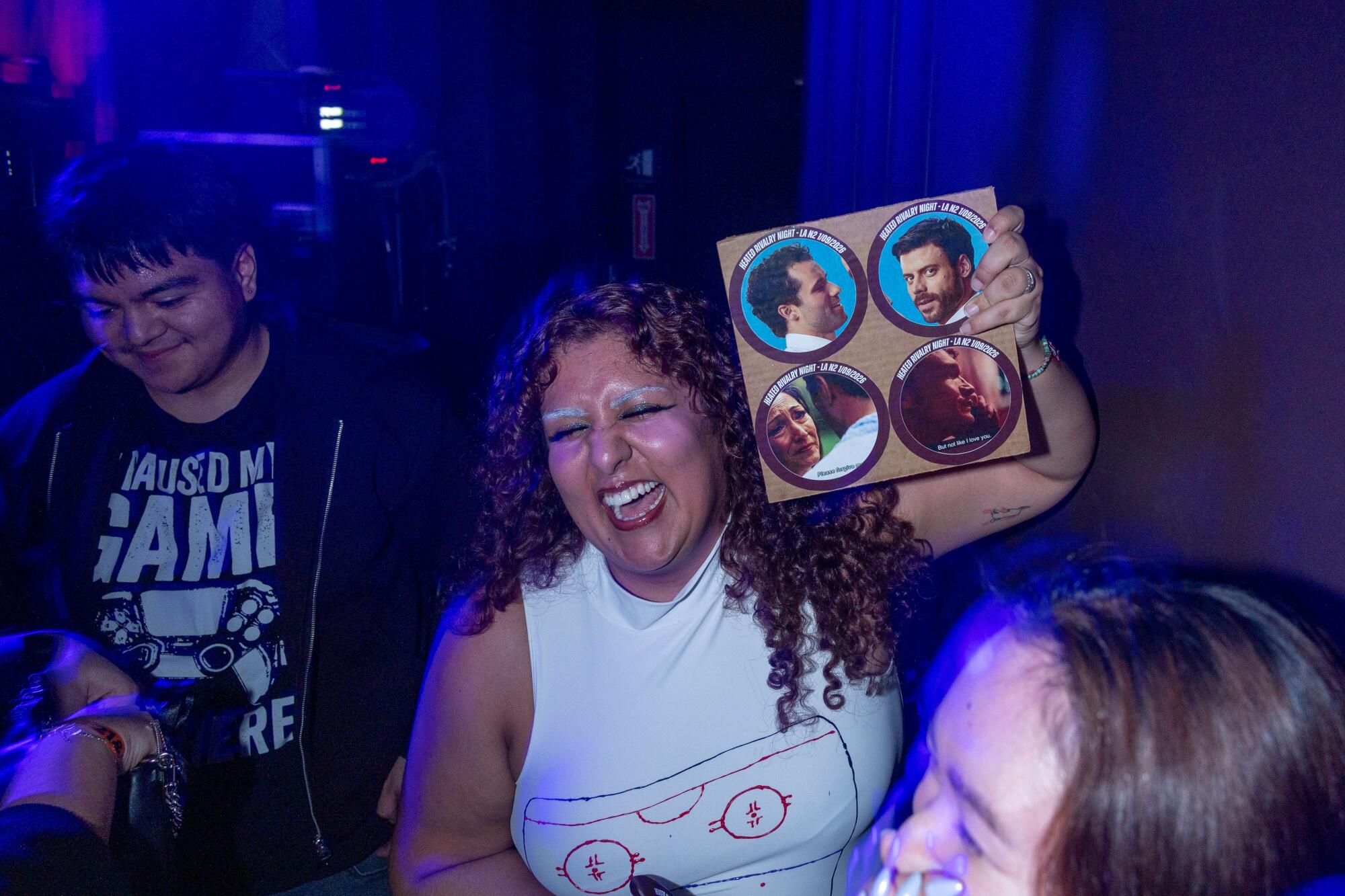 A woman in a white tank top holds up a small poster with circular cutouts of scenes from the TV series "Heated Rivalry."