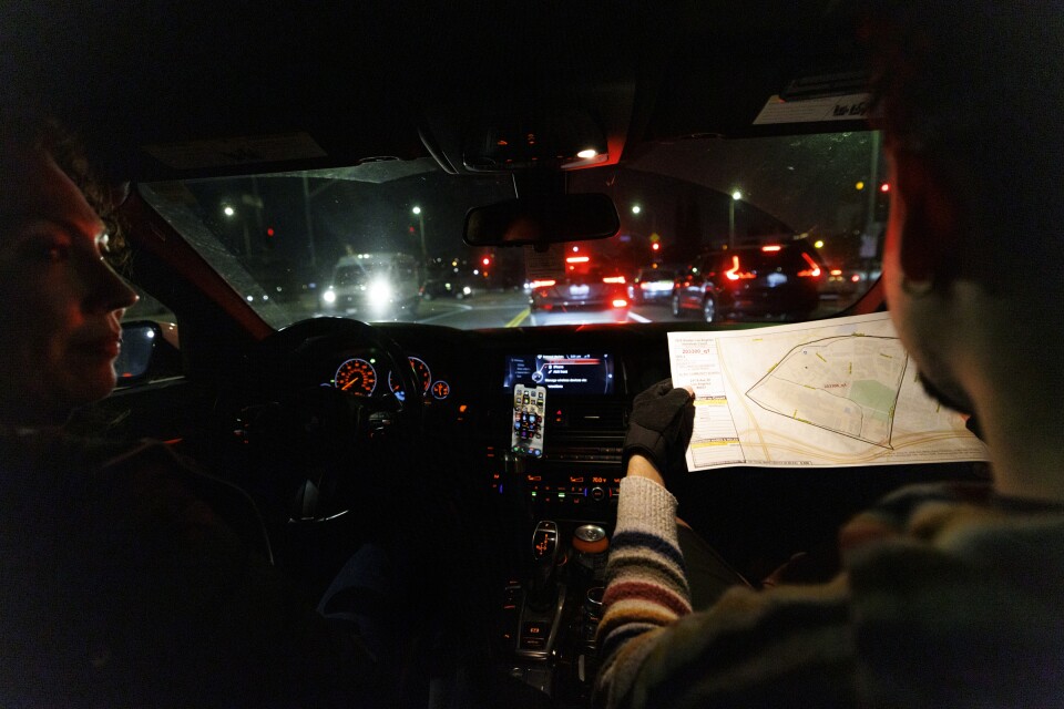 A woman driving in a car at night looks toward a man in the passenger seat holding out a map. 