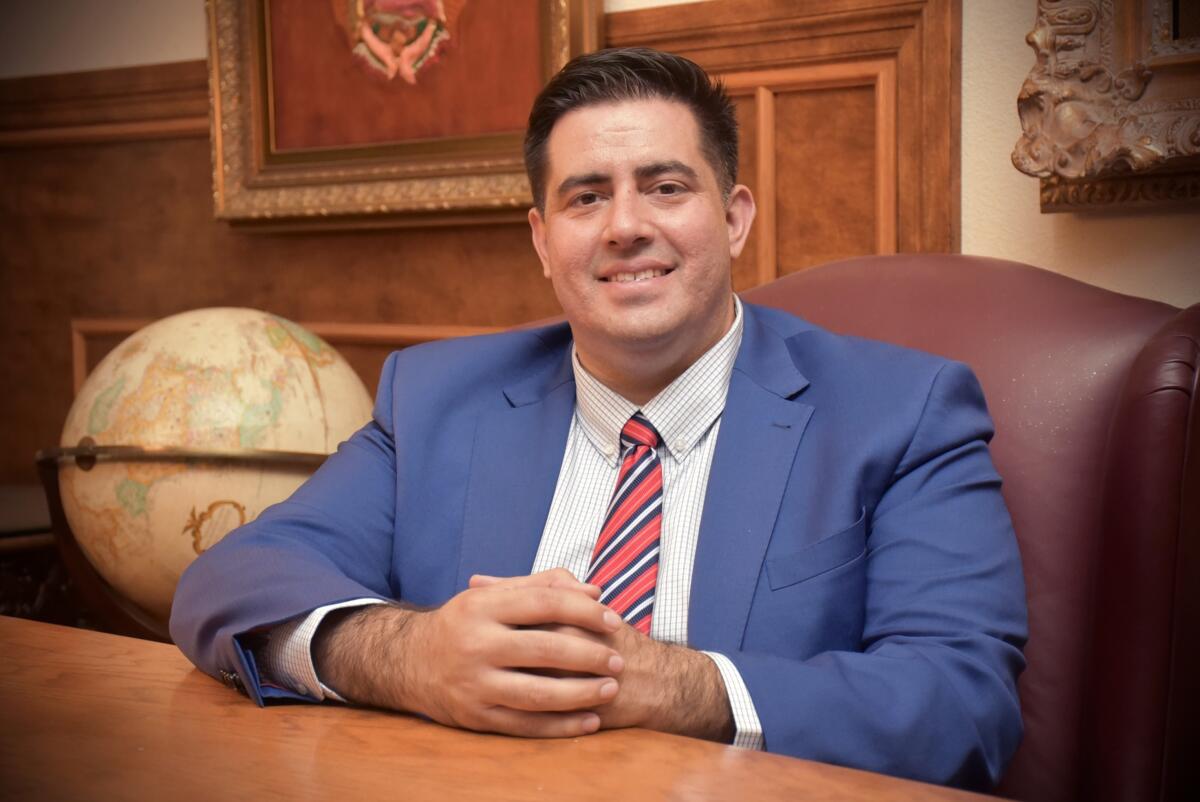 A man in a suite sits at a deck with his hands folded in front of him.