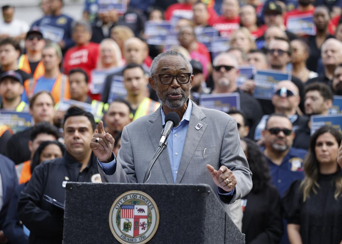 Councilmember Curren Price Jr. speaking at a lectern while several people stand behind him