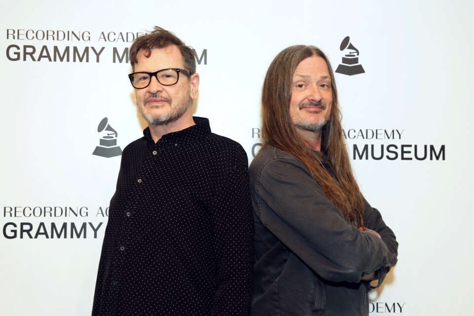 Two men, one with short hair and glasses and another with long hair and a mustache pose in front of a background that says "Grammy Museum."