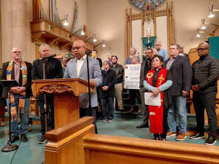 A man wearing a suit stands in front of a church pulpit and speaks into a microphone. Men and women wearing religious attire stand behind him. Once woman carries a sign that reads "Love not hate, makes America great."