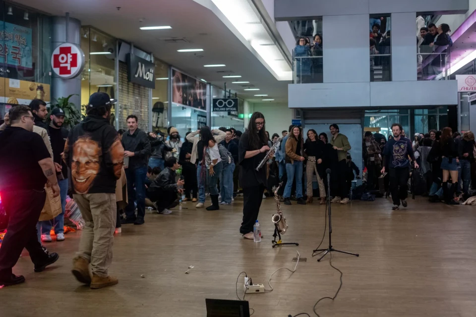 A person, wearing a black t-shirt and black pants, holds a clarinet next to a saxophone on a stand and microphone stand. There is a crowd of people around him watching and waiting in a mall.