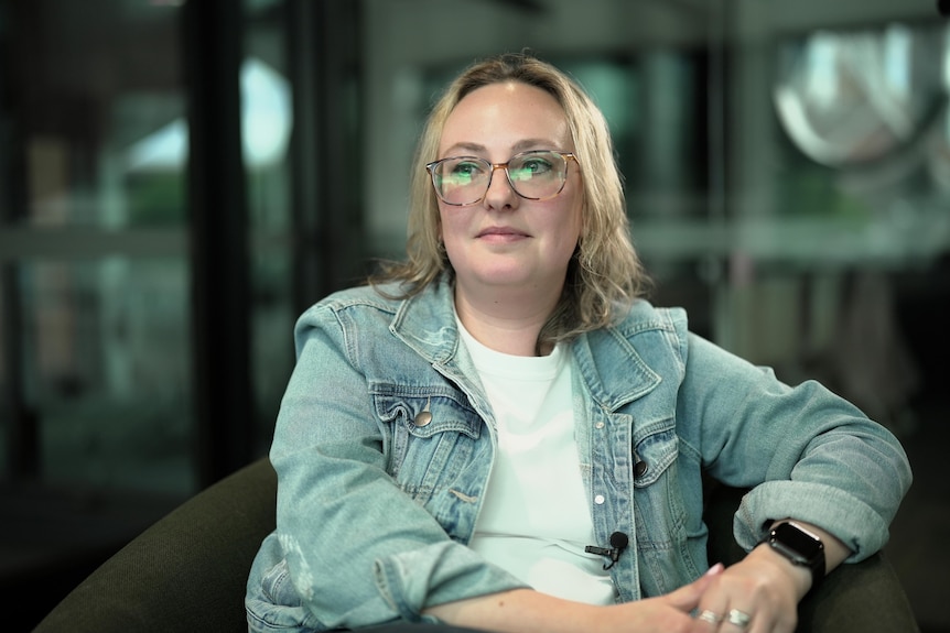 A woman with mid length blonde hair and glasses. She's wearing a light demin jacket and sitting in a chair
