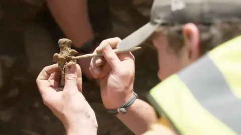 Kevin Church/BBC News Close-up of an archaeologist’s hands carefully cleaning a small bone fragment. They are holding the bone in one hand and using a thin wooden tool to scrape away the soil that it is covered with. The archaeologist is wearing a yellow high-visibility vest and a cap.