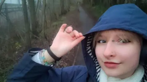 Amelka Zambrzycka A selfie image of Amelka from the shoulders up, she is wearing a navy rain coat with the hood up and a white scarf. She is on a gloomy walking path, it seems to have been raining and it's cold.