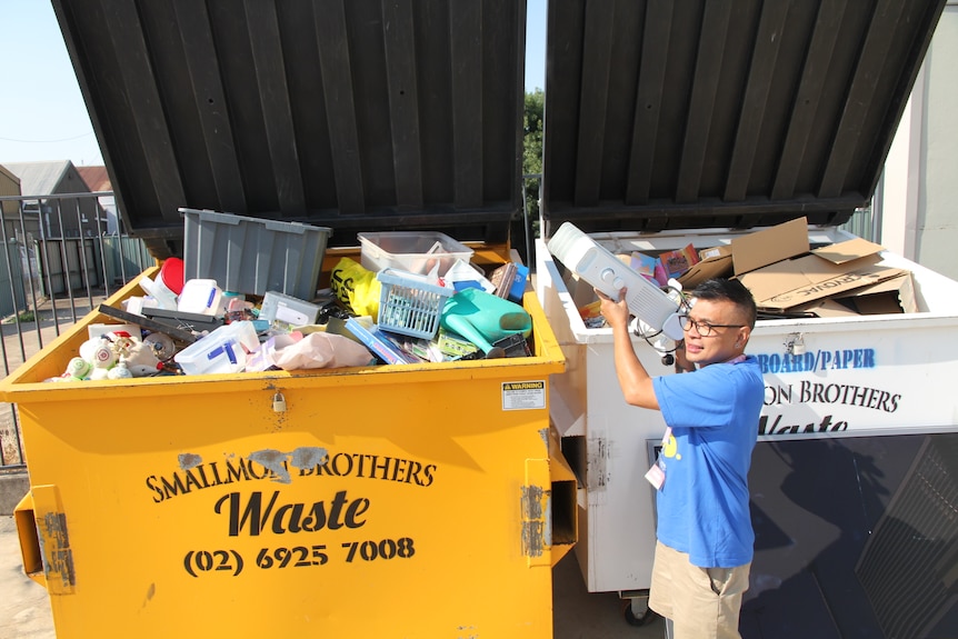 A man wearing a blue shirt and black hair throw an old oil heater into a skip bin which is overfilled