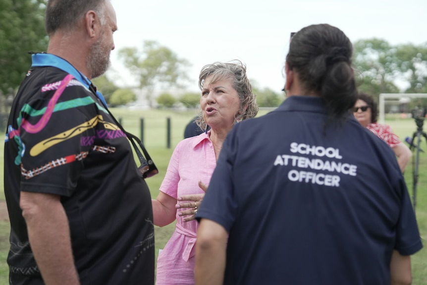 A woman in a pink dress speaking to people, one of them is wearing a shirt that reads: SCHOOL ATTENDANCE OFFICER