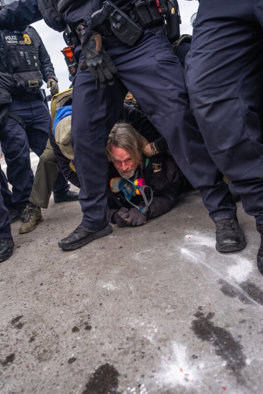 A person with long hair lies on the ground surrounded by several police officers in dark uniforms. The person appears to be detained, and their face is red. The scene is tense and takes place on a concrete surface.