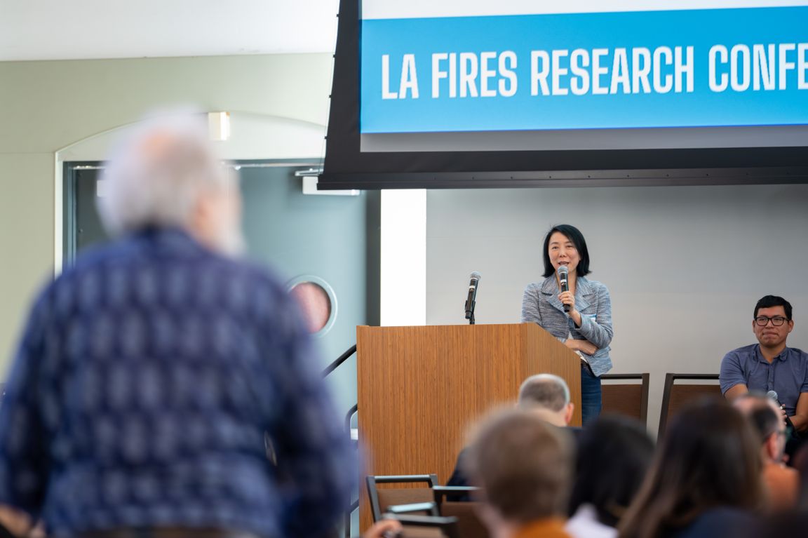 An audience member is shown with their back to the camera as they stand to address Yifang Zhu and other panelists in a Q&A session. 