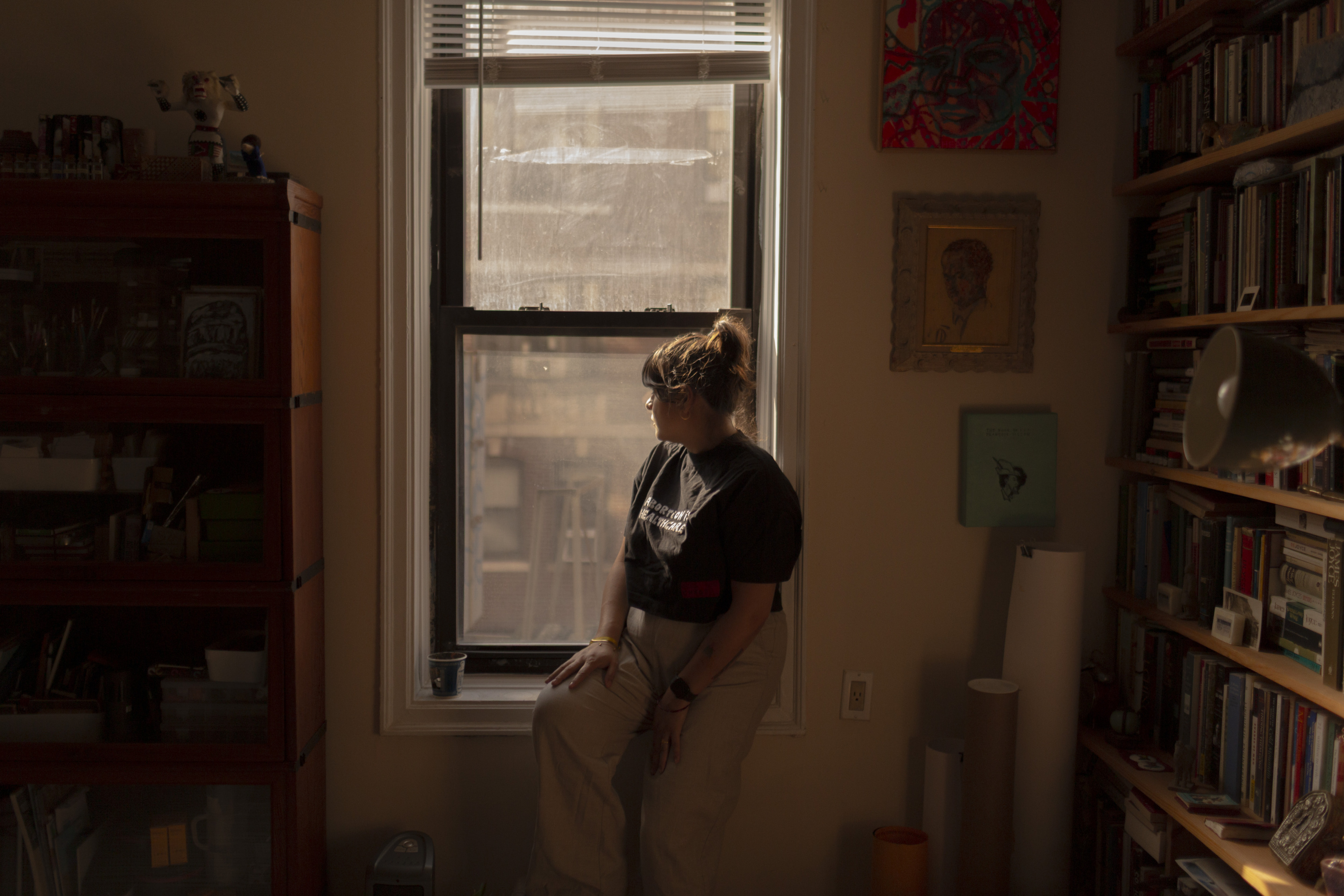 A woman facing away from the camera sits on a windowsill in a dimly lit room. She wears a black T-shirt and khaki pants and she is surrounded by bookcases filled with books and rolled up tubes of paper propped upright, with framed pictures hanging on the walls.
