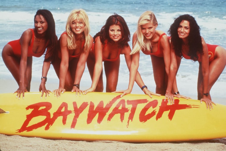 Actors pose with a large surfboard reading "Baywatch"