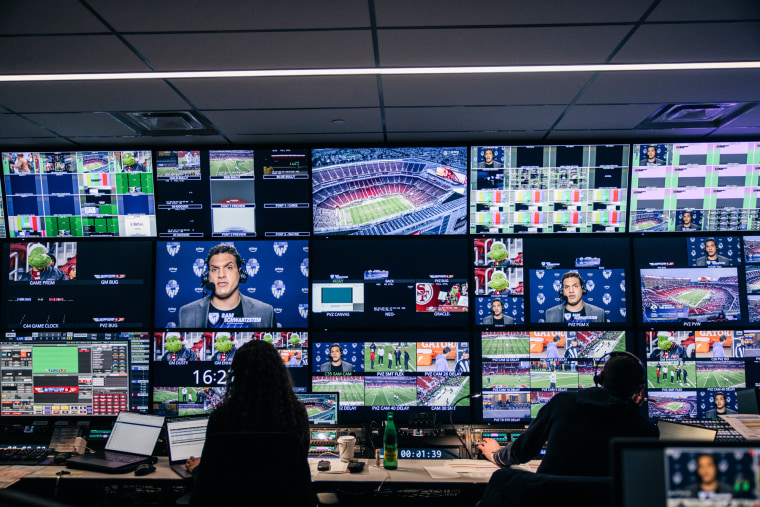 People sit in a control room in front of many computer and television screens displaying various angles of a football game