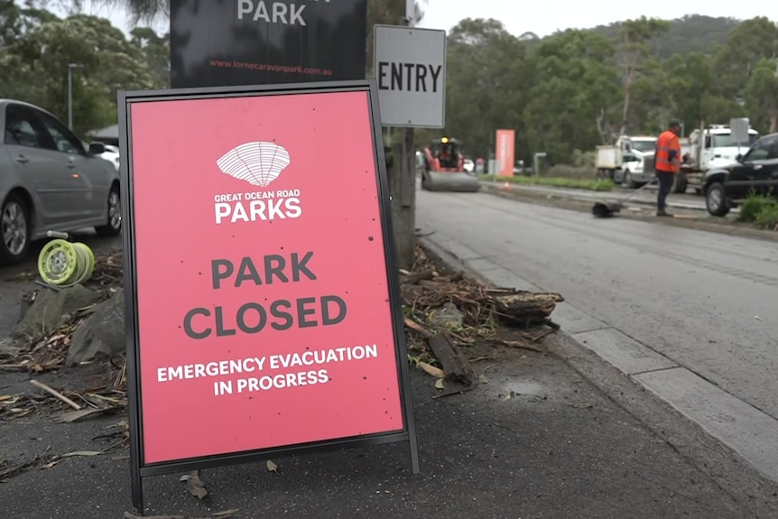 A red sign on a roadway says "park closed" and "emergency evacuation in progress" as a man in an orange shirt stands near a car.