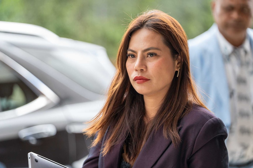 A young, dark-haired woman in a dark blazer stands near a vehicle parked on a city street.