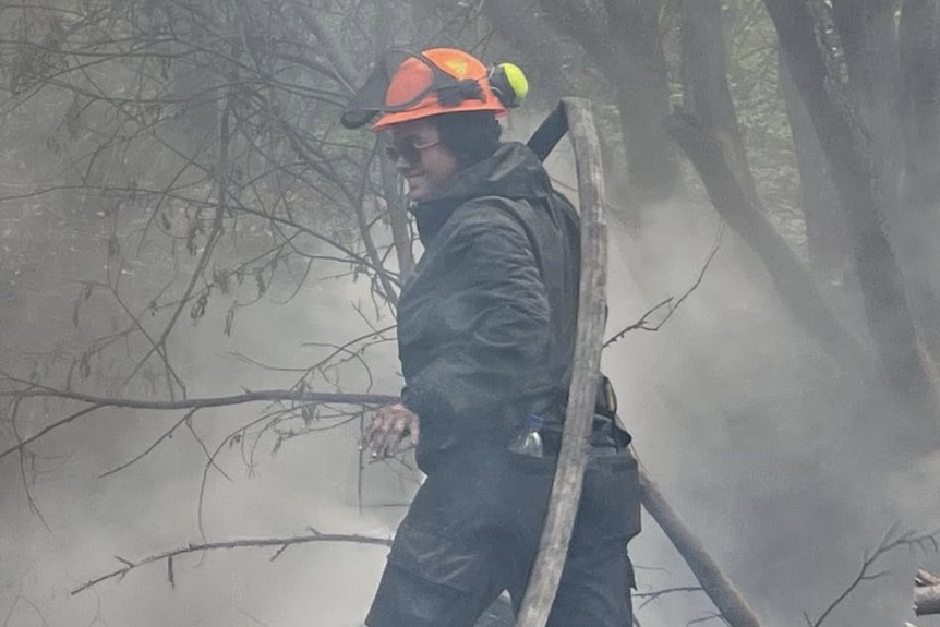 A woman in an orange hard hat and black fire-fighting clothes carrying a hose in a smokey area.